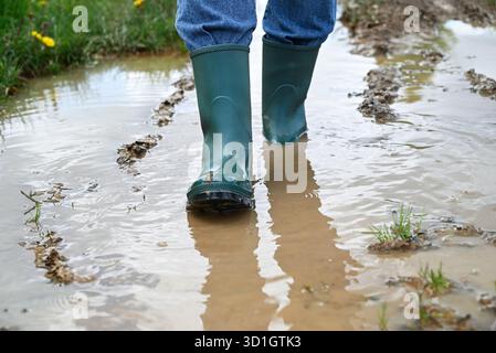 Person, die durch schlammige Pfützen auf einer ländlichen Feldstraße in grünen Gummistiefeln läuft. Stockfoto