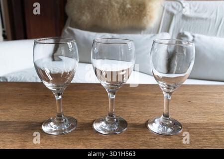 Drei identische, leere Weingläser stehen auf einem rustikalen Holztisch mit einem verschwommenen Hintergrund aus weißen Kissen und einem strukturierten Überwurf. Stockfoto