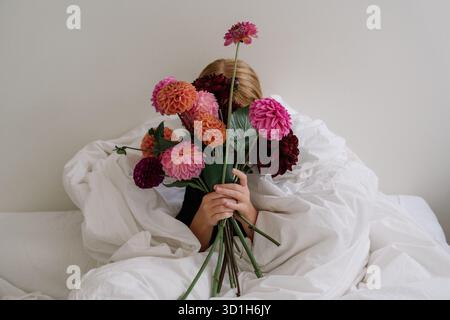 Ein Mädchen, das auf einem Bett sitzt, in eine Decke gewickelt und einen Strauß aus burgunderroten, rosa und orangen Dahlien hält. Minimalismus, Ästhetik Stockfoto