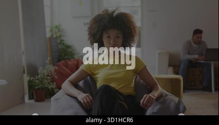 Sitzende Frau, die zu Hause ein gelbes T-Shirt auf einem Sitzsack-Stuhl trägt, während ein Freund sich mit einem Laptop zurücklehnt Stockfoto