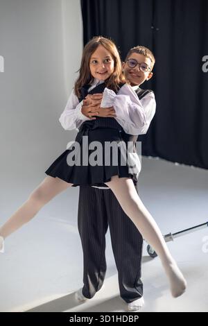 Ein fröhlicher Junge hebt ein Mädchen in verspielter Pose, in formeller Kleidung, und zeigt einen lustigen und energiegeladenen Moment in einem hellen Studio. Stockfoto
