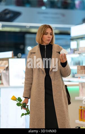 Beschreibung: Eine Frau, die ein weißes Kosmetikprodukt und eine gelbe Rose in der Hand hält, in einem hell beleuchteten Schönheits- oder Kosmetikgeschäft steht und das Produkt ansieht Stockfoto