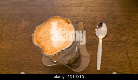Cremige Latte Art in einer Glasschale auf einem rustikalen Holztisch, begleitet von einem silbernen Löffel, die eine gemütliche Café-Atmosphäre vermittelt. Stockfoto