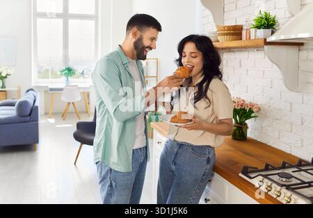 Romantisches Frühstück eines glücklichen Paares in der gemütlichen Küche zu Hause, Mann teilt Croissant Stockfoto
