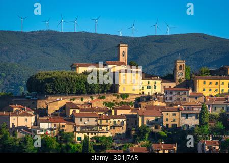 Blick auf das historische Dorf Montescudaio in der Toskana, Italien, mit der Kirche und dem Uhrenturm im Kontrast zu Windturbinen im Hintergrund. Stockfoto