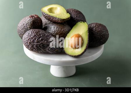 Reife Hass Avocados auf einem weißen Marmorständer, wobei einer offen geschnitten ist, um das grüne Fleisch und den Samen vor einem weichen grünen Hintergrund zu zeigen. Stockfoto