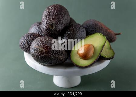 Reife Hass Avocados auf einem weißen Marmorständer, wobei einer offen geschnitten ist, um das grüne Fleisch und den Samen vor einem weichen grünen Hintergrund zu zeigen. Stockfoto