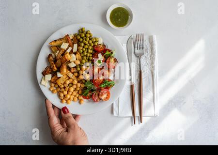 Blick von oben auf eine Frau, die nach einem Mittagstisch mit Tomaten- und Koriandersalat, Kichererbsen, grünen Erbsen und gebratenen Kartoffeln mit Käse und einer scharfen Sauce greift Stockfoto