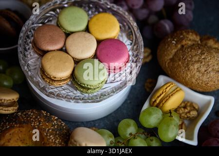 Eine kleine Schüssel mit Macarons sitzt zwischen Trauben und Brötchen auf einer rustikalen Tischplatte. Der Overhead-Winkel zeigt Vielfalt und Farbe an, wodurch ein Versati entsteht Stockfoto