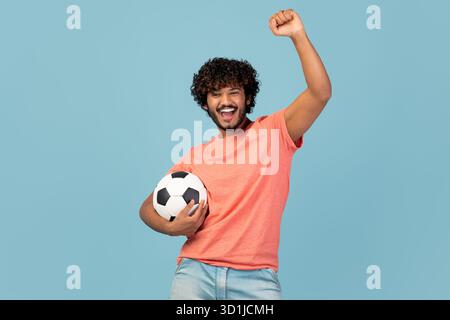 Junger indischer Mann, der als Fußballfan in einem blauen Studio-Hintergrund feiert Stockfoto