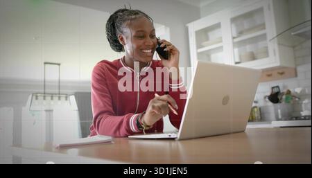 Sprechende Frau in rotem Sweatshirt mit Smartphone und Schreiben auf Laptop in der Küche, Notizbuch daneben Stockfoto