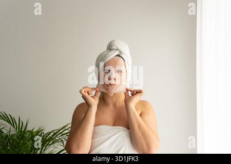 Schöne Frau, die weiße Gesichtsmaske mit Handtuch auf Kopf nach Dusche im Spa-Badezimmer mit grüner Pflanze für Hautpflege Schönheitsroutine anwendet Stockfoto