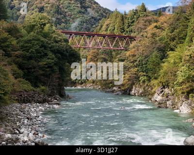 Eine Brücke überspannt einen Fluss mit einem grünen und blauen Wasserweg. Die Brücke ist rot und erstreckt sich über die gesamte Breite des Flusses. Die Brücke ist von Bäumen und umgeben Stockfoto