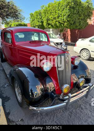 Oldtimer in Rot und Schwarz parkt auf einer sonnendurchfluteten Straße mit Bäumen und anderen Fahrzeugen im Hintergrund Stockfoto