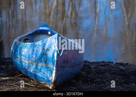 Verwittertes blaues Kanu an der Muddy Shoreline mit Wasserreflexionen im Hintergrund, bereit für Abenteuer Stockfoto
