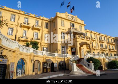 Historische gelbe Fassade des Winter Palace Hotels mit fliegenden Fahnen und geschwungener Treppe unter klarem blauem Himmel Stockfoto