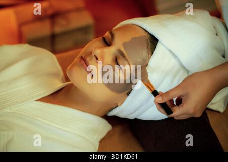 In der ruhigen Atmosphäre des Spa-Salons verwöhnt die Kundin ihre Gäste mit einer Gesichtscreme mit Holzkohle und einer warmen Kerze. Gesichtshaut Stockfoto