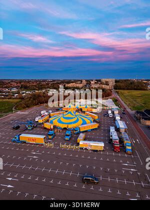 Blick auf den Sonnenuntergang von Circus Renz, gelbblaues Zelt umgeben von Lastwagen und Anhängern auf einem geräumigen Parkplatz, lebhaftem Himmel und nahe gelegenen Häusern Stockfoto