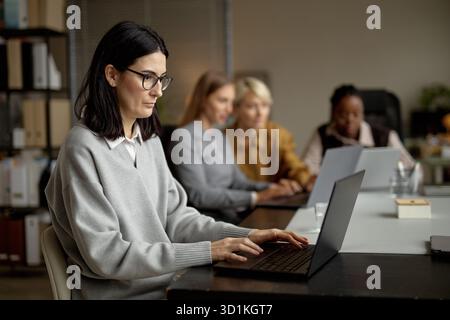 Erwachsene kaukasische Frau, die an einem Laptop arbeitet, mit verschiedenen Kollegen, die zusammenarbeiten Stockfoto