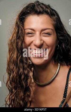 Eine fröhliche junge Frau mit lockigen Haarstrahlen vor Glück, die ihr helles Lächeln zeigt. Sie trägt eine stilvolle Halskette, die Wärme und Positivität ausstrahlt Stockfoto