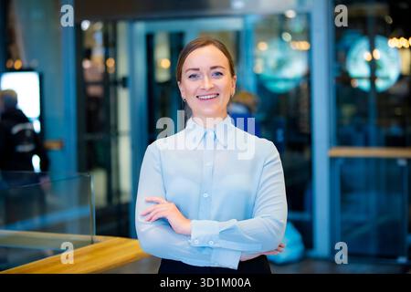 Selbstbewusste Geschäftsfrau mittleren Alters lächelt in der modernen Glasbürohalle in die Kamera, professionelles Firmenporträt mit natürlichem Licht. Stockfoto