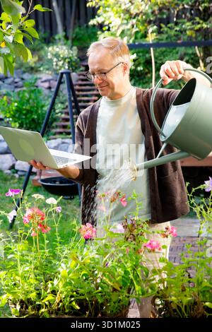 Mann mittleren Alters, der Blumen gießt und während eines Videoanrufs auf der sonnigen Terrasse fernarbeitet, modernes Multitasking-Lifestyle-Konzept. Stockfoto