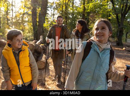 Vierköpfige Familie spaziert durch den sonnigen Wald, genießt ihre Wanderung, während sie Wanderstöcke trägt und tagsüber zusammen lächelt Stockfoto