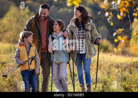 Eine vierköpfige Familie spaziert durch die sonnige Wiese, genießt ihre Wanderung, während sie Wanderstöcke trägt und tagsüber zusammen lächelt Stockfoto