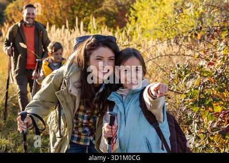 Eine vierköpfige Familie spaziert durch die sonnige Wiese, genießt ihre Wanderung, während sie Wanderstöcke trägt und tagsüber zusammen lächelt Stockfoto