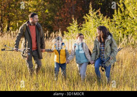 Eine vierköpfige Familie spaziert durch die sonnige Wiese, genießt ihre Wanderung, während sie Wanderstöcke trägt und tagsüber zusammen lächelt Stockfoto