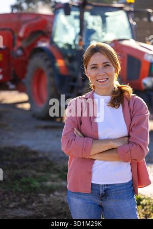 Porträt einer jungen Bäuerin, die mit gekreuzten Armen vor dem Traktor mit Futtermischer auf der Rinderfarm steht Stockfoto
