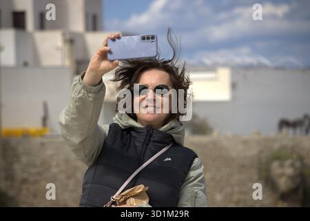 Eine lächelnde Frau, die eine Sonnenbrille und eine zweifarbige Pufferjacke trägt, hält ein Smartphone in der Hand, um ein Selfie zu machen. Ihr Haar wird vom Wind zerzaust Stockfoto