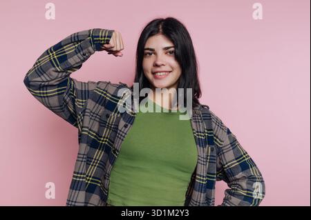 Eine junge Frau in einem karierten Hemd, die ihren Arm beugt und selbstbewusst vor einem rosa Hintergrund lächelt. Stockfoto
