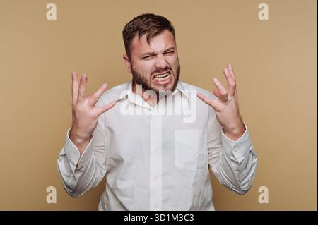Ein frustrierter Mann in einem weißen Hemd, der Wut vor beigefarbenem Hintergrund ausdrückt. Stockfoto