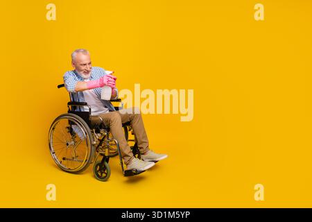 Fröhlicher Opa in einer Rollstuhlreinigung mit Sprühflasche vor hellgelbem Hintergrund, der die Hoffnung auf die Reha und die Einbeziehung in den Alltag zelebriert Stockfoto