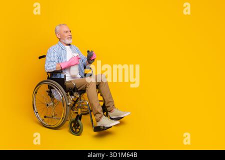 Lustiger Winkel Opa in einem hellen Rollstuhl genießt einen Reha-Moment mit pinkfarbenen Handschuhen vor einem auffälligen gelben Hintergrund Stockfoto