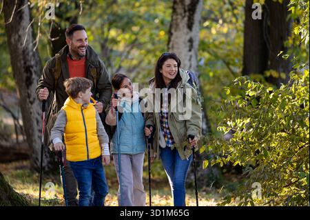 Vierköpfige Familie spaziert durch den sonnigen Wald, genießt ihre Wanderung, während sie Wanderstöcke trägt und tagsüber zusammen lächelt Stockfoto