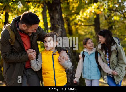 Vierköpfige Familie spaziert durch den sonnigen Wald, genießt ihre Wanderung, während sie Wanderstöcke trägt und tagsüber zusammen lächelt Stockfoto