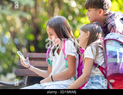 Fröhliche Schulkinder sitzen in der Pause mit ihren Rucksäcken auf einer Bank und schauen sich lustige Videos auf einem digitalen Gerät an Stockfoto