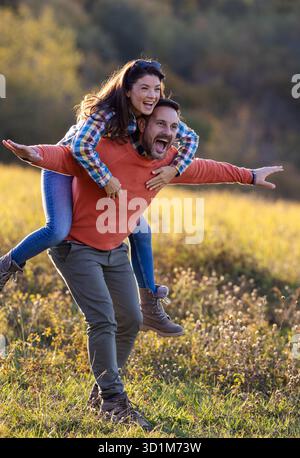 Ein glückliches Paar hat tagsüber Spaß auf einem grasbewachsenen Feld. Der Mann trägt die Frau auf dem Rücken, während sie lächeln und spielerisch posieren Stockfoto