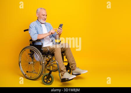 Lustiger Seniorengroßvater in einem Rollstuhl verwendet ein Smartphone vor einem hellgelben Hintergrund für ein warmes inklusive Stockbild über Gesundheit und Stockfoto
