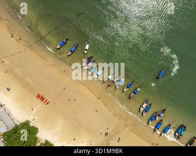 Blick aus der Vogelperspektive auf Longtail-Boote, die am Ufer liegen, während türkisfarbene Wellen sanft gegen den goldenen Sand, Ao Nang, Krabi, Thailand, schlagen. Stockfoto