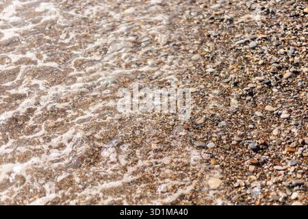 Die Küste ist mit glatten Kieselsteinen und Sand bedeckt, während sanfte Wellen an Land strömen. Sonnenlicht schimmert auf dem Wasser und hebt die Farben der Steine hervor. Stockfoto