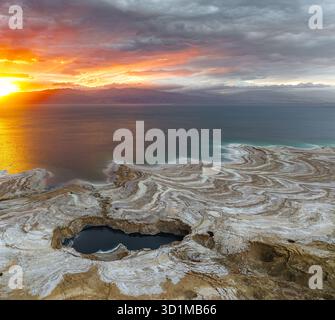 Aus der Vogelperspektive auf die faszinierende Küste des Toten Meeres, wo der lebhafte Sonnenuntergang den Himmel übermalt, während er auf die einzigartigen Salzformationen trifft, ein Gedi, Israel. Stockfoto