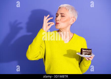 Ältere Frau im hellgelben Pullover genießt Kuchen und einen verspielten Kuss vor einem lila Hintergrund in einem fröhlichen Lifestyle-Moment Stockfoto