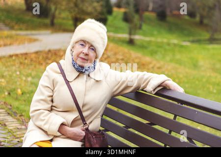 Ernsthafte Seniorin sitzt im Herbstpark mit warmer Jacke und Schal. Ältere Frau, die in die Kamera blickt, mit ruhigem Ausdruck, natürlichen Outdoor-Portren Stockfoto