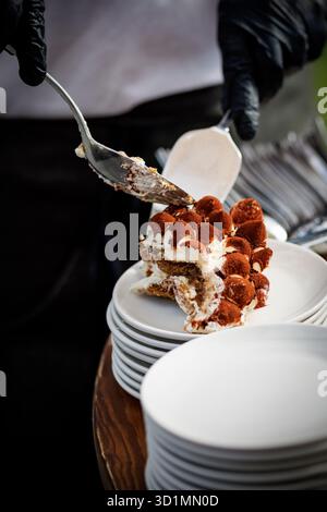 Ein Koch mit schwarzen Handschuhen serviert ein Stück Tiramisu-Kuchen auf gestapelten weißen Tellern bei einer Veranstaltung. Stockfoto