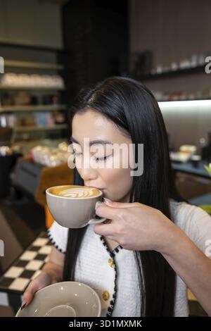 Taillenbildnis einer jungen asiatischen Frau, die Kaffee mit Schaum aus der Keramikschale trinkt Stockfoto