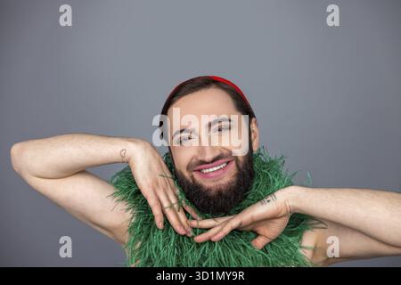 Nahaufnahme Porträt einer lächelnden, zufriedenen, dunkelhaarigen transsexuellen Person, die einen grünen Boa-Schal trägt, hält die Hände unter dem Kinn isoliert über grauem Hinterkopf Stockfoto
