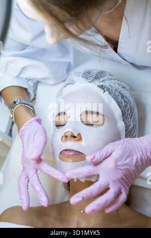 Unerkennbarer Kosmetologe mit Gummihandschuhen, der eine Blattmaske auf das Gesicht der Frau aufträgt Stockfoto